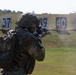 A soldier in full combat gear, including a tactical helmet and multicam camouflage uniform with a backpack, is firing a rifle on a military shooting range. Numbered target boards (36, 37, 39, 40, 41) are visible downrange on a grassy field. A spent casing appears to be ejecting from the weapon, captured mid-flight.