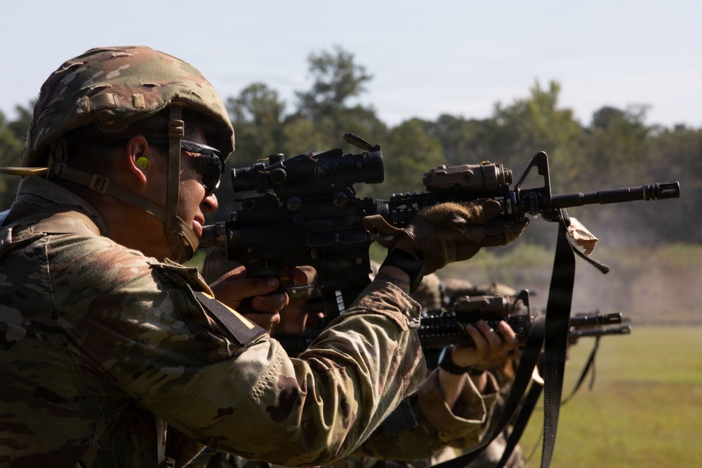Two soldiers in MultiCam/OCP camouflage uniforms are engaged in a live-fire shooting exercise on an outdoor range. The foreground soldier is aiming an M4/M16-style carbine equipped with an optic sight and additional rail-mounted accessories. Smoke is visible in the background, consistent with active firing.
