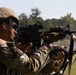 Two soldiers in MultiCam/OCP camouflage uniforms are engaged in a live-fire shooting exercise on an outdoor range. The foreground soldier is aiming an M4/M16-style carbine equipped with an optic sight and additional rail-mounted accessories. Smoke is visible in the background, consistent with active firing.