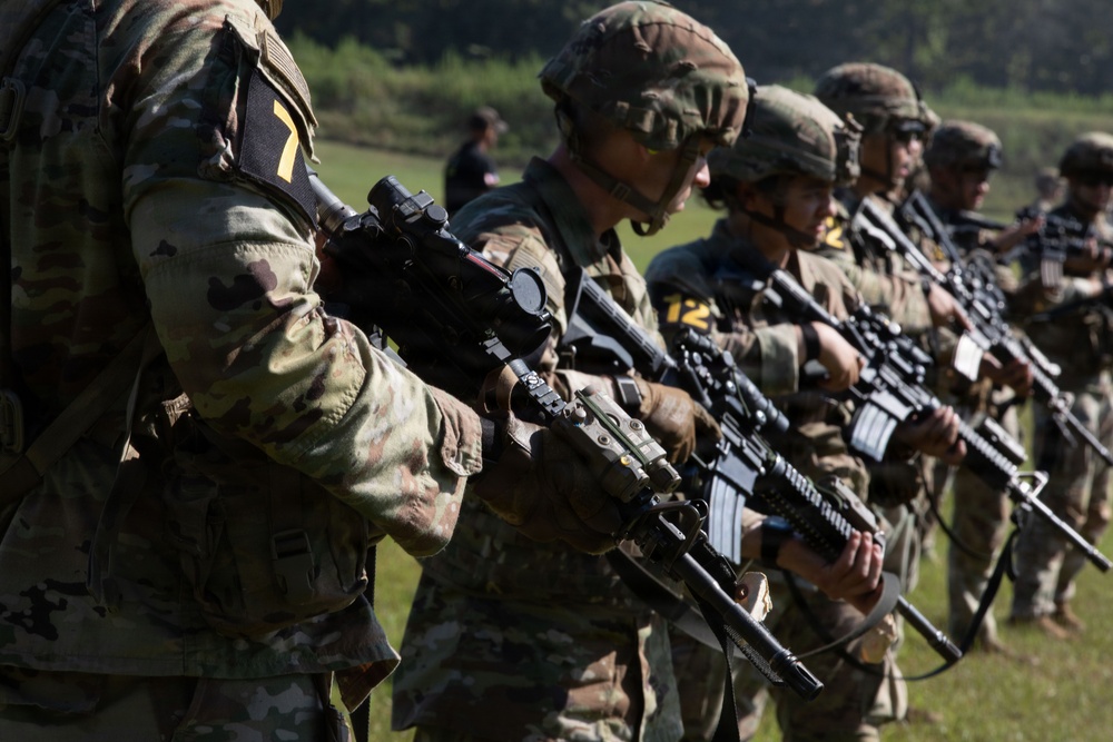 A line of military personnel in full combat gear are standing in formation holding rifles, likely during a training exercise or inspection. They are wearing MultiCam/OCP camouflage uniforms with tactical helmets and equipment. Numbered placards (visible numbers 7 and 12) are attached to their gear, suggesting a competitive or evaluation event.