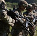 A line of military personnel in full combat gear are standing in formation holding rifles, likely during a training exercise or inspection. They are wearing MultiCam/OCP camouflage uniforms with tactical helmets and equipment. Numbered placards (visible numbers 7 and 12) are attached to their gear, suggesting a competitive or evaluation event.