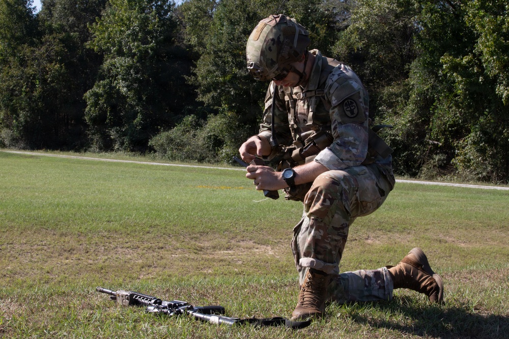 A soldier in full combat gear is kneeling on a grass field, appearing to handle or manipulate equipment with both hands. An assault rifle (appears to be an M4/M16-style weapon) is laid on the ground in front of them. The setting appears to be a military training area bordered by trees.
