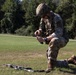 A soldier in full combat gear is kneeling on a grass field, appearing to handle or manipulate equipment with both hands. An assault rifle (appears to be an M4/M16-style weapon) is laid on the ground in front of them. The setting appears to be a military training area bordered by trees.