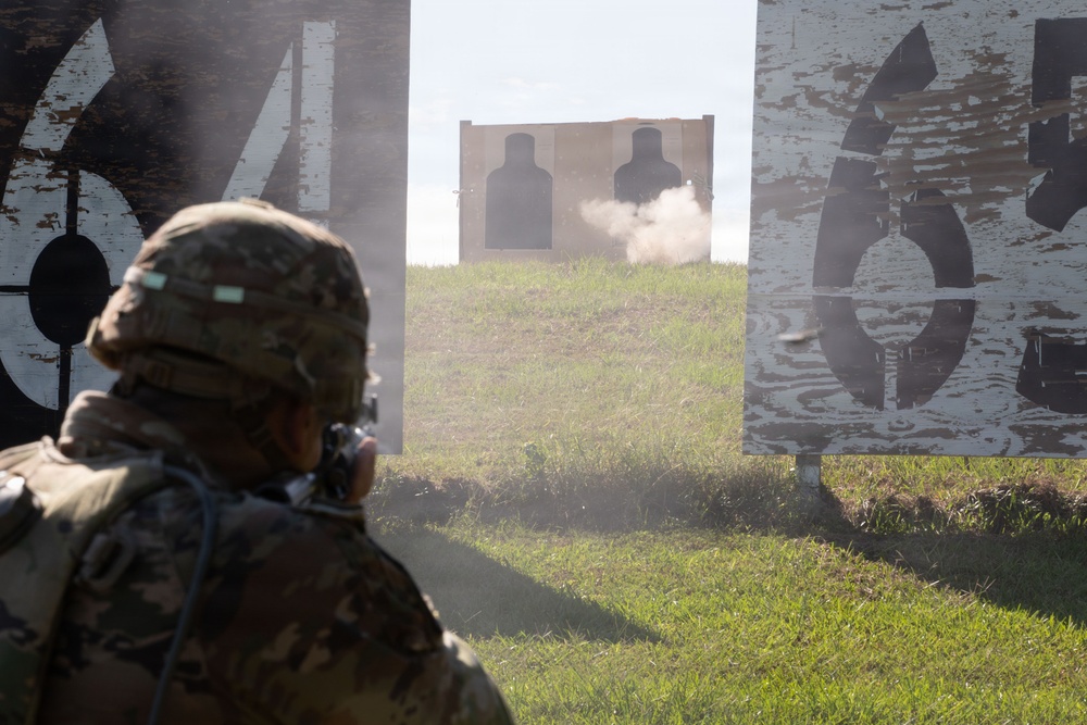 A soldier in full combat gear and helmet is engaged in a live-fire training exercise at a shooting range. The soldier is aiming at two silhouette targets downrange, with visible smoke/dust indicating a recent or active shot. Large numbered range markers are visible on either side of the lane.