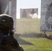 A soldier in full combat gear and helmet is engaged in a live-fire training exercise at a shooting range. The soldier is aiming at two silhouette targets downrange, with visible smoke/dust indicating a recent or active shot. Large numbered range markers are visible on either side of the lane.