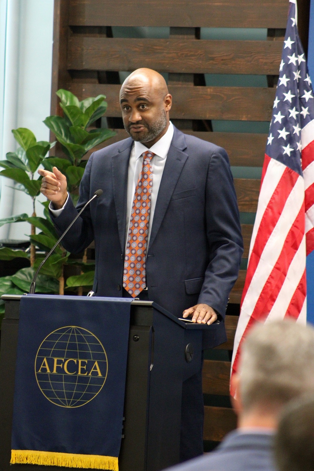 A man in a navy blue suit and orange patterned tie is speaking at a podium bearing the AFCEA (Armed Forces Communications and Electronics Association) logo. He is gesturing with his right hand while addressing an audience, with an American flag visible to his right. The setting appears to be a formal indoor event or conference.