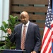 A man in a navy blue suit and orange patterned tie is speaking at a podium bearing the AFCEA (Armed Forces Communications and Electronics Association) logo. He is gesturing with his right hand while addressing an audience, with an American flag visible to his right. The setting appears to be a formal indoor event or conference.