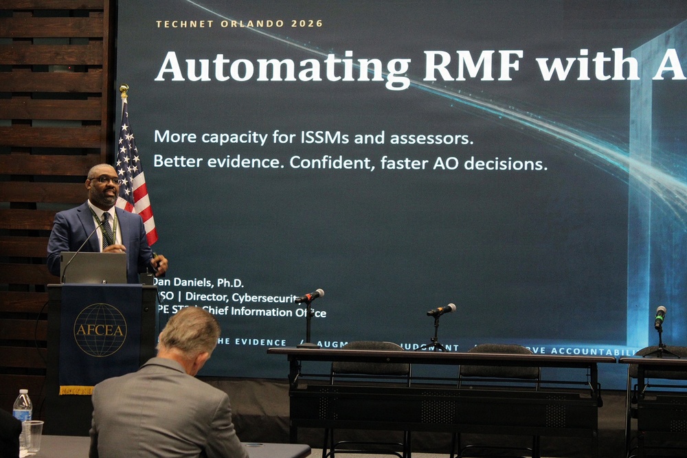 A presenter identified as Dan Daniels, Ph.D., ISO and Director of Cybersecurity at a Chief Information Office, is delivering a talk at TechNet Orlando 2026 hosted by AFCEA. The presentation is titled 'Automating RMF with A[I]' and focuses on improving capacity for ISSMs, assessors, and enabling faster AO decisions. An audience member is visible in the foreground seated with their back to the camera.
