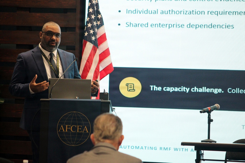A man in a navy blue suit is presenting at an AFCEA (Armed Forces Communications and Electronics Association) event, standing at a podium with a laptop. Behind him, a projected presentation slide discusses topics including 'individual authorization requirements,' 'shared enterprise dependencies,' and 'the capacity challenge' related to 'Automating RMF with AI.' An American flag is visible in the background.