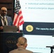 A man in a navy blue suit is presenting at an AFCEA (Armed Forces Communications and Electronics Association) event, standing at a podium with a laptop. Behind him, a projected presentation slide discusses topics including 'individual authorization requirements,' 'shared enterprise dependencies,' and 'the capacity challenge' related to 'Automating RMF with AI.' An American flag is visible in the background.