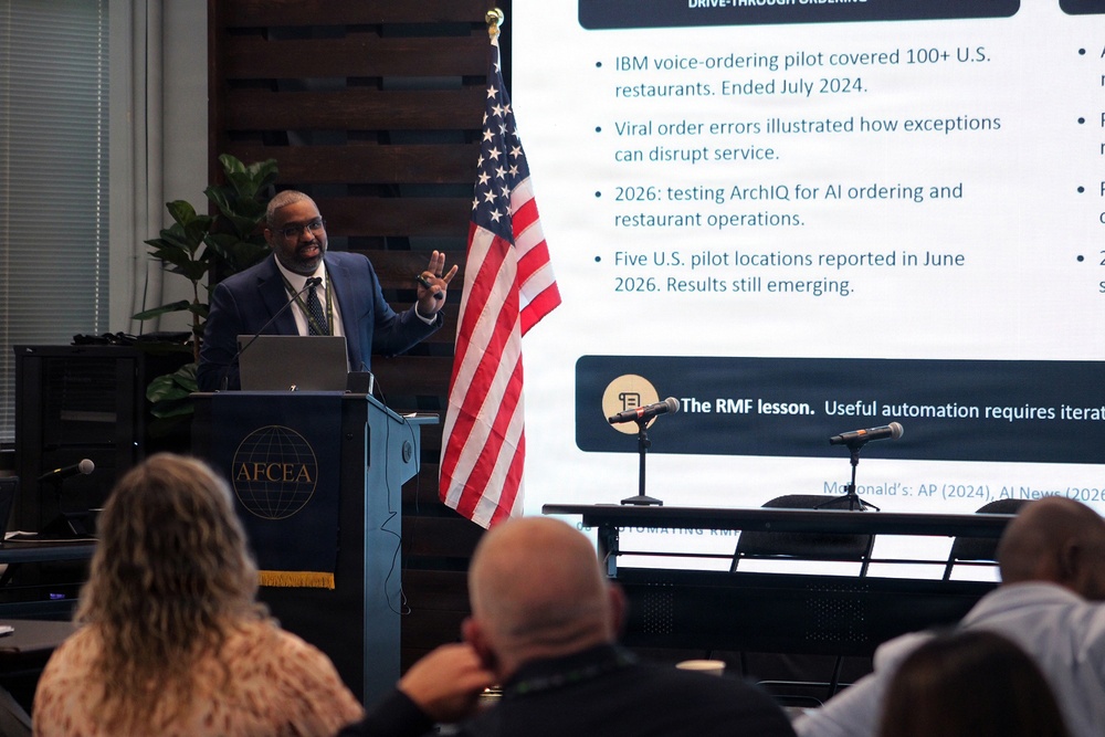 A man in a blue suit is presenting at an AFCEA podium before an audience, with a large projection screen behind him displaying content about AI drive-through ordering systems, including references to IBM voice-ordering pilots and McDonald's. The presentation slide discusses topics such as viral order errors, ArchIQ AI testing, and a highlighted 'RMF lesson' about automation requiring iteration. An American flag stands to the right of the speaker.
