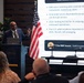 A man in a blue suit is presenting at an AFCEA podium before an audience, with a large projection screen behind him displaying content about AI drive-through ordering systems, including references to IBM voice-ordering pilots and McDonald's. The presentation slide discusses topics such as viral order errors, ArchIQ AI testing, and a highlighted 'RMF lesson' about automation requiring iteration. An American flag stands to the right of the speaker.