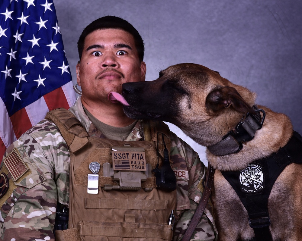 A U.S. Air Force Staff Sergeant named PITA, assigned to KILO 9 / TSGB unit, poses in a formal military portrait with his Military Working Dog (MWD). The dog is licking the handler's face, creating a candid and humorous moment during what appears to be an official photo session. An American flag is visible in the background, consistent with standard military portrait settings.