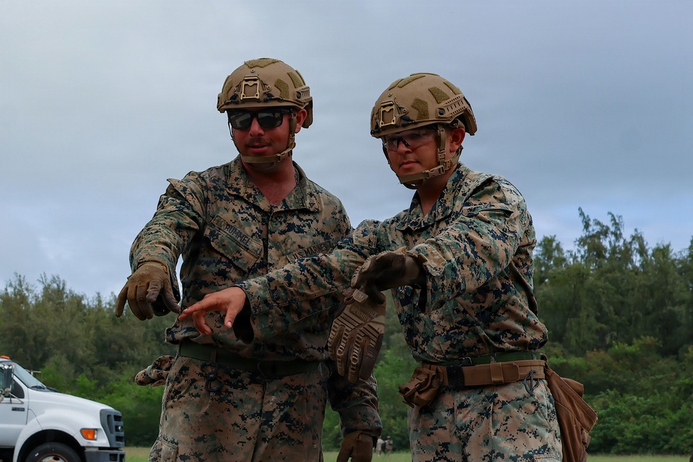 Two U.S. Marines in full combat gear are engaged in what appears to be a briefing or coordination discussion in an outdoor field environment. Both wear MARPAT digital camouflage uniforms, tactical helmets, and protective eyewear. One Marine appears to be gesturing or pointing, possibly explaining a task or procedure to the other.