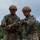 Two U.S. Marines in full combat gear are engaged in what appears to be a briefing or coordination discussion in an outdoor field environment. Both wear MARPAT digital camouflage uniforms, tactical helmets, and protective eyewear. One Marine appears to be gesturing or pointing, possibly explaining a task or procedure to the other.