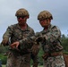 Two U.S. Marines in full combat gear are engaged in what appears to be a briefing or coordination discussion in an outdoor field environment. Both wear MARPAT digital camouflage uniforms, tactical helmets, and protective eyewear. One Marine appears to be gesturing or pointing, possibly explaining a task or procedure to the other.