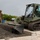 A military construction scene showing a U.S. Marine in combat uniform observing a JCB compact track loader painted in olive drab military green moving gravel on a concrete surface. The equipment and personnel appear to be engaged in construction or repair work at a military installation. The setting appears to be a tropical or subtropical location based on the vegetation visible in the background.