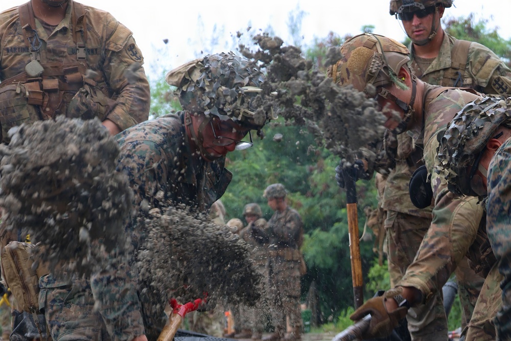 Military personnel in mixed uniforms — including USMC MARPAT and US Army OCP/Multicam — are actively digging or working with soil/dirt, with debris visibly flying through the air. The scene appears to be a field training exercise involving entrenching or demolition-related ground work. Multiple service members are engaged simultaneously, suggesting a coordinated team task.