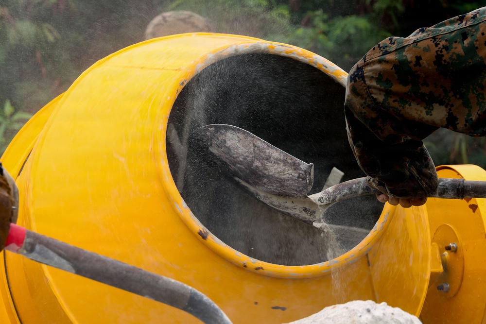 A worker wearing camouflage clothing is shoveling material, likely cement or sand, into a yellow rotating concrete mixer drum. Dust and particles are visibly airborne around the opening of the mixer, indicating active loading. The scene appears to be an outdoor construction site with vegetation visible in the background.
