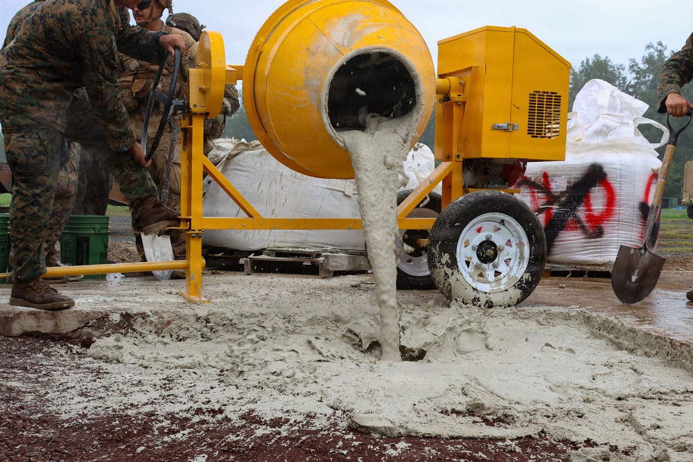 Military personnel in camouflage uniforms are operating a yellow portable concrete mixer, pouring wet concrete onto a prepared ground surface. The scene appears to be a field construction or engineering operation, likely a military training or infrastructure project. Large bags of dry concrete mix are visible behind the mixer, and workers with shovels are managing the pour.