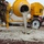 Military personnel in camouflage uniforms are operating a yellow portable concrete mixer, pouring wet concrete onto a prepared ground surface. The scene appears to be a field construction or engineering operation, likely a military training or infrastructure project. Large bags of dry concrete mix are visible behind the mixer, and workers with shovels are managing the pour.