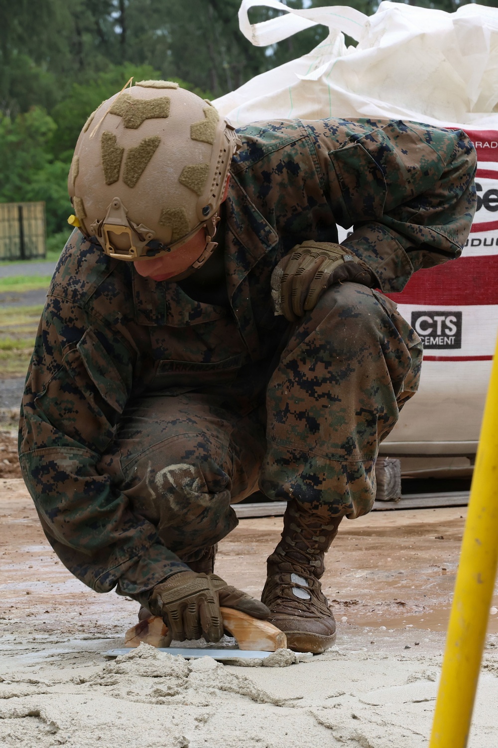 A military service member in MARPAT (Marine digital camouflage) uniform is crouched down working on a concrete or cement surface, using a trowel or finishing tool. The individual is wearing full combat gear including a tan FAST helmet with velcro patches, tactical gloves, and combat boots. A large bag of CTS Cement is visible in the background, indicating a construction or repair task.