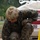 A military service member in MARPAT (Marine digital camouflage) uniform is crouched down working on a concrete or cement surface, using a trowel or finishing tool. The individual is wearing full combat gear including a tan FAST helmet with velcro patches, tactical gloves, and combat boots. A large bag of CTS Cement is visible in the background, indicating a construction or repair task.