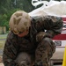 A military service member in MARPAT (Marine digital camouflage) uniform is crouched down working on a concrete or cement surface, using a trowel or finishing tool. The individual is wearing full combat gear including a tan FAST helmet with velcro patches, tactical gloves, and combat boots. A large bag of CTS Cement is visible in the background, indicating a construction or repair task.