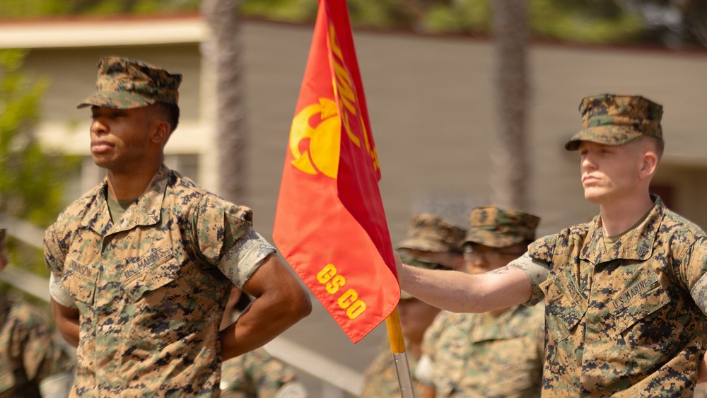 Two U.S. Marines in MARPAT woodland camouflage uniforms stand at attention during a formal military ceremony. One Marine holds a red guidon flag bearing the Eagle, Globe, and Anchor emblem with the designation 'GS CO' (General Support Company). Additional Marines are visible in the background, suggesting a unit formation or parade.