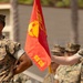 Two U.S. Marines in MARPAT woodland camouflage uniforms stand at attention during a formal military ceremony. One Marine holds a red guidon flag bearing the Eagle, Globe, and Anchor emblem with the designation 'GS CO' (General Support Company). Additional Marines are visible in the background, suggesting a unit formation or parade.
