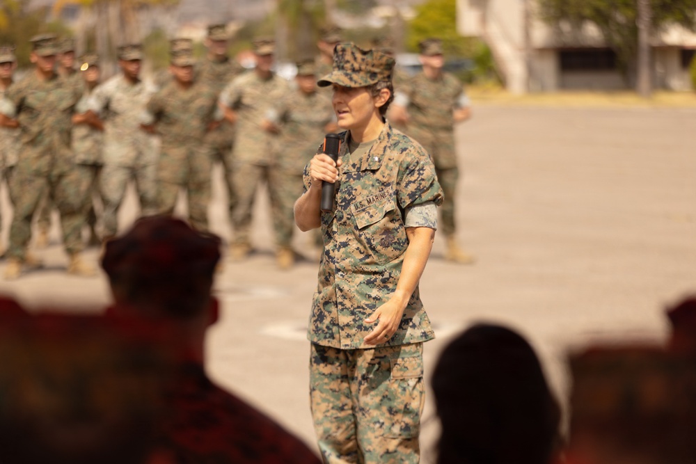 A U.S. Marine in MARPAT camouflage uniform is addressing a formation while holding a microphone, likely during a ceremony or briefing on a military installation. A large group of Marines in uniform stands in formation in the background. The setting appears to be an outdoor parade deck or assembly area.