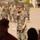 A U.S. Marine in MARPAT camouflage uniform is addressing a formation while holding a microphone, likely during a ceremony or briefing on a military installation. A large group of Marines in uniform stands in formation in the background. The setting appears to be an outdoor parade deck or assembly area.