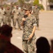 A U.S. Marine in MARPAT camouflage uniform is addressing a formation while holding a microphone, likely during a ceremony or briefing on a military installation. A large group of Marines in uniform stands in formation in the background. The setting appears to be an outdoor parade deck or assembly area.