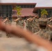 A formation of military personnel in camouflage uniforms are executing a salute during what appears to be a formal ceremony or parade. The scene takes place on a paved parade ground in front of a building with a red roof, with palm trees visible in the background. The image is shot from a low angle through the foreground figures, creating a depth-of-field effect.