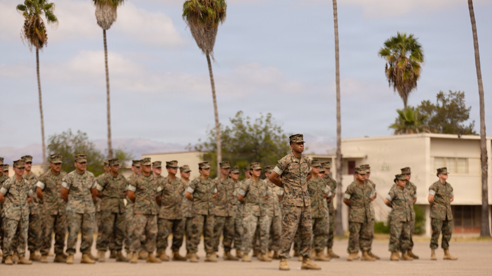 A formation of military personnel in camouflage uniforms stands at parade rest on an outdoor drill pad. One individual, likely a non-commissioned officer or drill instructor, stands facing the formation in a position of authority. The setting appears to be a military base in a warm, arid climate based on the palm trees and desert terrain visible in the background.