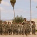 A formation of military personnel in camouflage uniforms stands at parade rest on an outdoor drill pad. One individual, likely a non-commissioned officer or drill instructor, stands facing the formation in a position of authority. The setting appears to be a military base in a warm, arid climate based on the palm trees and desert terrain visible in the background.
