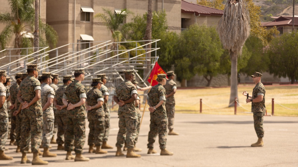 A formation of U.S. Marines in MARPAT camouflage uniforms stands at parade rest on a drill deck while a single Marine addresses them from the front. A red unit guidon flag is visible within the formation. The setting appears to be a military installation with barracks buildings, bleachers, palm trees, and arid landscaping visible in the background.