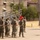 A formation of U.S. Marines in MARPAT camouflage uniforms stands at parade rest on a drill deck while a single Marine addresses them from the front. A red unit guidon flag is visible within the formation. The setting appears to be a military installation with barracks buildings, bleachers, palm trees, and arid landscaping visible in the background.