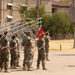A formation of U.S. Marines in MARPAT camouflage uniforms stands at parade rest on a drill deck while a single Marine addresses them from the front. A red unit guidon flag is visible within the formation. The setting appears to be a military installation with barracks buildings, bleachers, palm trees, and arid landscaping visible in the background.
