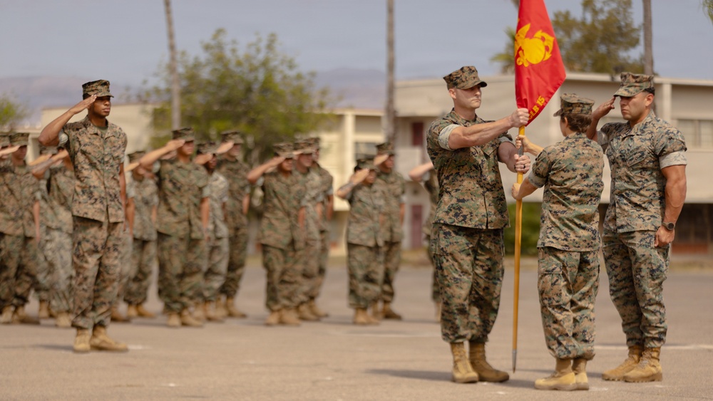 A U.S. Marine Corps military ceremony is taking place on a parade deck, likely a change of command or unit activation ceremony. Personnel in MARPAT camouflage uniforms are standing in formation and rendering salutes while a guidon (unit flag) is being passed between two Marines in the foreground. The setting appears to be a military installation with barracks-style buildings visible in the background.
