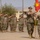 A U.S. Marine Corps military ceremony is taking place on a parade deck, likely a change of command or unit activation ceremony. Personnel in MARPAT camouflage uniforms are standing in formation and rendering salutes while a guidon (unit flag) is being passed between two Marines in the foreground. The setting appears to be a military installation with barracks-style buildings visible in the background.