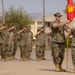 A U.S. Marine Corps military ceremony is taking place on a parade deck, likely a change of command or unit activation ceremony. Personnel in MARPAT camouflage uniforms are standing in formation and rendering salutes while a guidon (unit flag) is being passed between two Marines in the foreground. The setting appears to be a military installation with barracks-style buildings visible in the background.