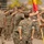 A U.S. Marine Corps military ceremony is taking place outdoors, likely a change of command or unit formation event. Personnel in MARPAT (Marine Pattern) camouflage uniforms are saluting while central figures appear to be transferring a unit guidon (flag). The setting appears to be a parade deck or drill field, consistent with a formal military installation.