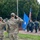 A military ceremony is taking place outdoors, featuring a color guard presenting flags while personnel in camouflage uniforms render salutes. The ceremony appears to be a formal military honors event, likely at a memorial or installation. A U.S. Air Force flag and American flag are prominently displayed by honor guard members in dress blues.
