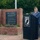 An elderly man in a blue suit with a red poppy pin and a veterans organization cap speaks at a podium during a POW/MIA memorial ceremony. Behind him stands a brick monument engraved with the 'Code of Conduct,' and an American flag is prominently displayed to his right. The setting appears to be an outdoor memorial garden.