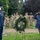 A military memorial ceremony is taking place outdoors in a park-like setting. Two individuals in the foreground — one in Air Force dress blues and one in a civilian suit with a veteran's cap — are rendering salutes before a floral wreath on a stand. A formation of soldiers in Army combat uniforms stands at attention in the background.