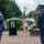 A formal military memorial ceremony is taking place at a POW/MIA memorial site. A veteran in a blue suit and garrison cap is rendering a salute while a uniformed Air Force service member stands at attention beside a white floral wreath. A third military figure stands at a podium bearing the POW/MIA emblem, with an American flag displayed nearby.