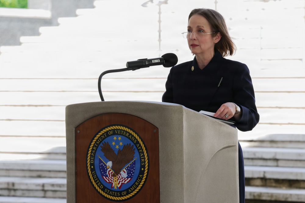 A woman in a dark navy jacket is speaking at a podium bearing the official seal of the Department of Veterans Affairs, United States of America. She appears to be delivering a formal address at an outdoor ceremony, likely at a memorial or government facility. The setting features marble or stone steps in the background, suggesting a significant civic or commemorative venue.