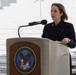 A woman in a dark navy jacket is speaking at a podium bearing the official seal of the Department of Veterans Affairs, United States of America. She appears to be delivering a formal address at an outdoor ceremony, likely at a memorial or government facility. The setting features marble or stone steps in the background, suggesting a significant civic or commemorative venue.