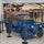 A blue U.S. Air Force military utility vehicle (M151 MUTT jeep) is on display inside what appears to be a military exchange store (AAFES). The vehicle is roped off with blue stanchions and bears markings identifying it as a McChord Air Museum exhibit. An American flag stands to the left, and Halloween decorations labeled 'The Boo Crew' are visible in the store background.